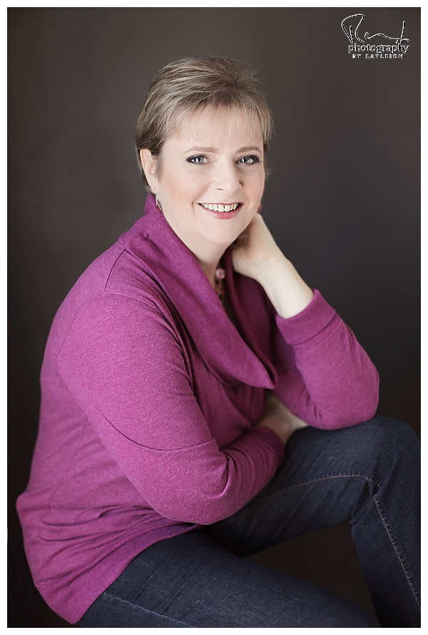 This image is a professional business headshot. The woman is in a sitting pose in front of a black background, wearing jeans and a sweater. Photography by Rayleigh. Byrayleigh.com