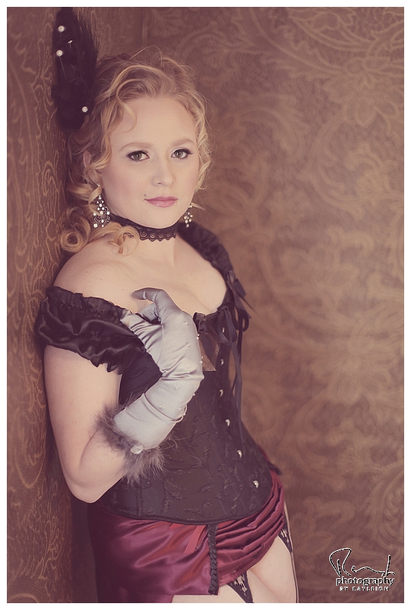Victorian inspired boudoir session with a wild west saloon girl theme. This is an image of a stylized vintage themed boudoir standing pose. She is wearing a black corset, choker necklace, and feather in her hair in front of a brown background. For more info, please visit byrayleigh.com