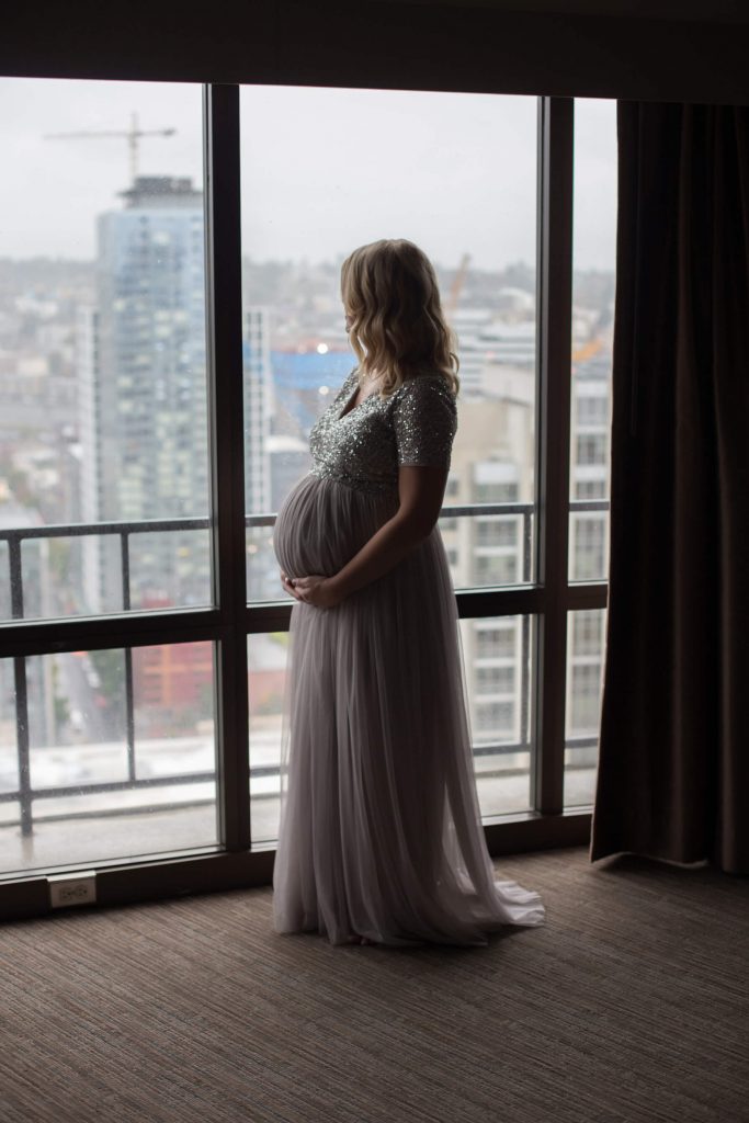 What to wear for a maternity photo shoot in Portland, OR. This is an image of a pregnant woman standing in front of a hotel room window with backlighting. She is dressed in a formal gown.