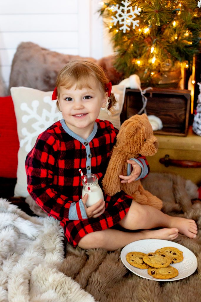 Christmas pajama sessions. This is an image of a little girl wearing red plaid Christmas pajamas. She's holding a glass of milk and a teddy bear, next to a plate of cookies, and sitting in front of a Christmas tree. Image by Photography by Rayleigh. For more info, visit byrayleigh.com