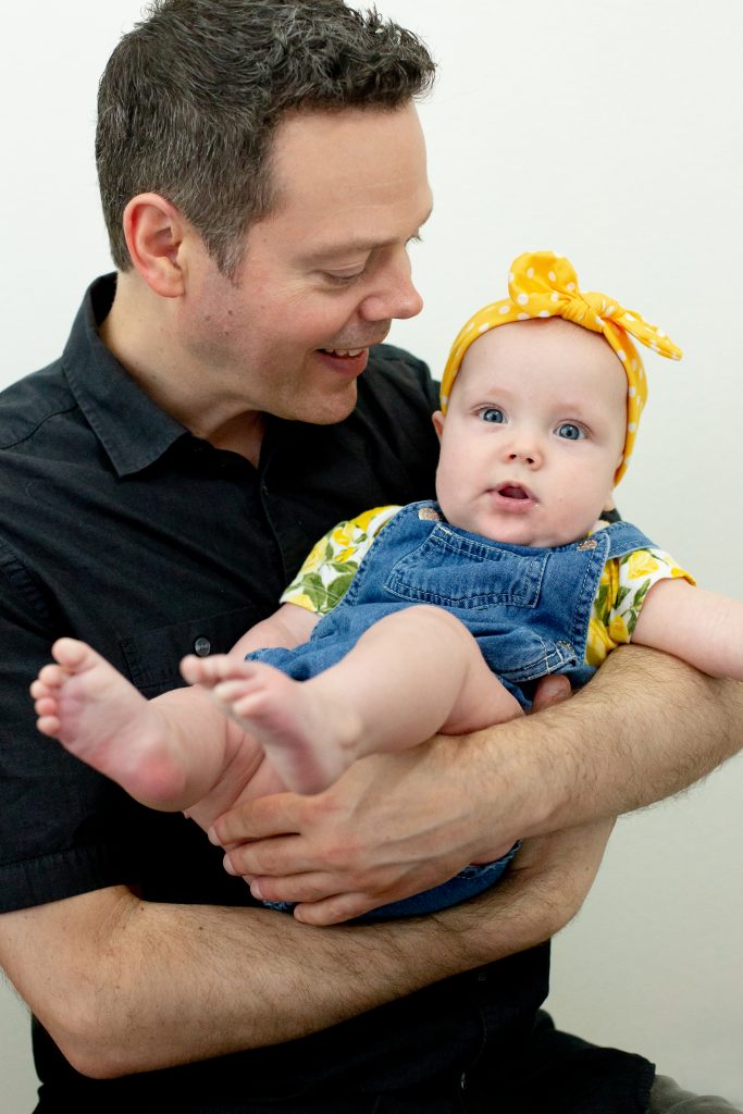 Father's Day gift ideas. This family photo was taken with natural light in a studio with a father holding his baby daughter and smiling in front of a white background. He is wearing a black polo shirt she is wearing yellow floral shirt with overalls and a yellow polka dot headband. This photo was taken by Photography by Rayleigh in Portland, OR. See more at byrayleigh.com.
