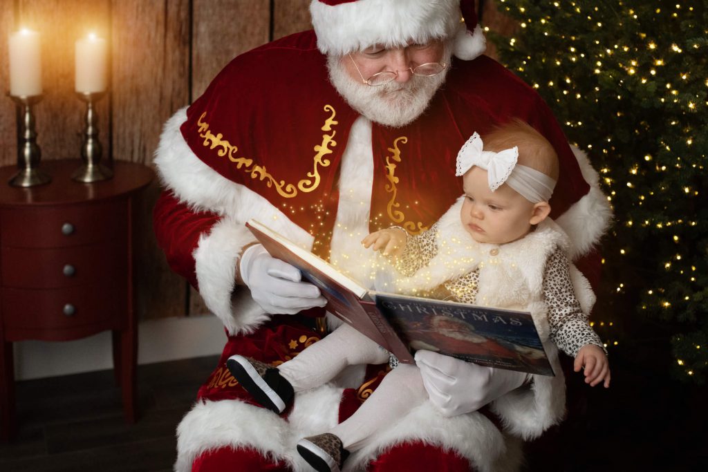 Santa Sessions in Portland, OR. Experience the magic of Christmas. Santa is reading a book to the baby girl during their photo shoot.