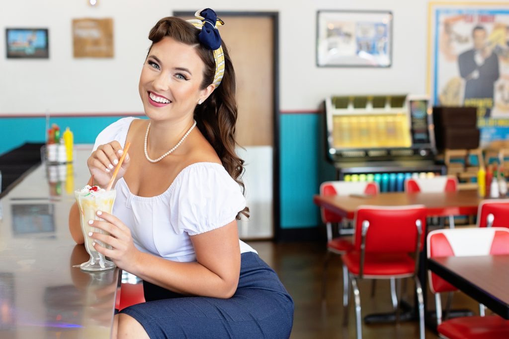 A pin up inspired diner photo shoot in Portland, OR by Photography by Rayleigh. This is a retro themed image of a smiling woman sitting at a diner bar, with a milkshake.