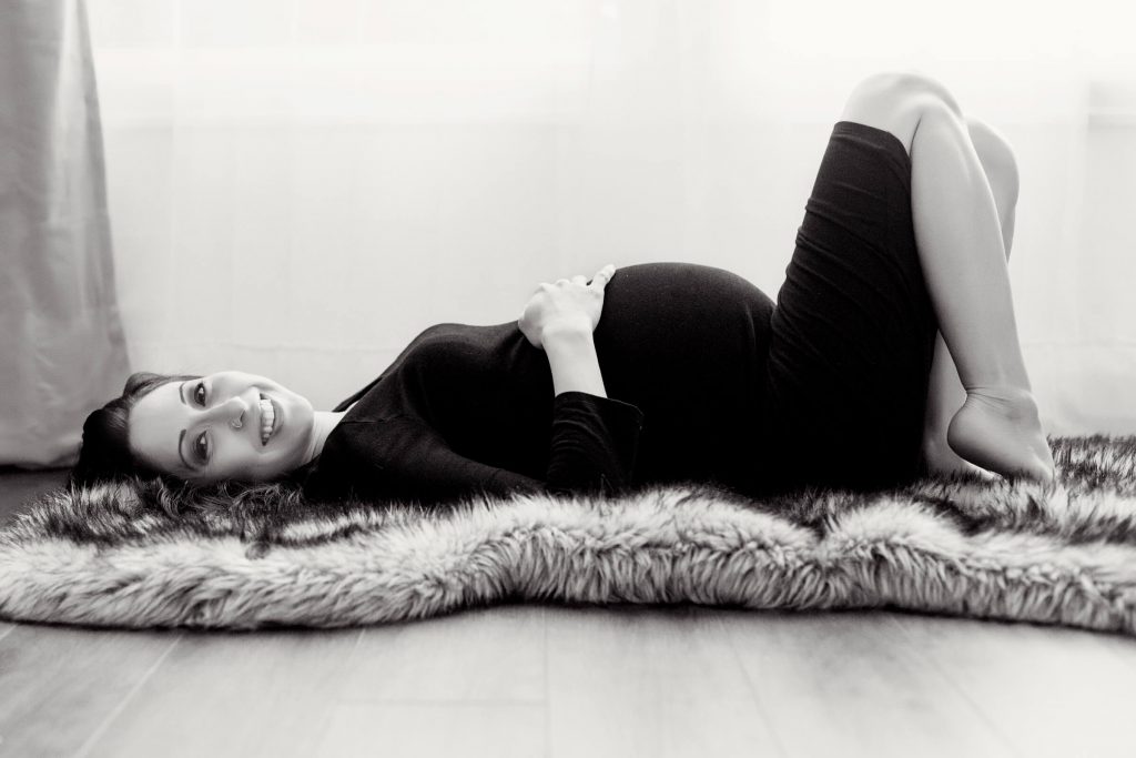 What to wear for a maternity photo shoot in Portland, OR. This is a black and white maternity boudoir image of a pregnant woman, in a floor pose, lying on her back on a fur rug in a black form fitted dress.