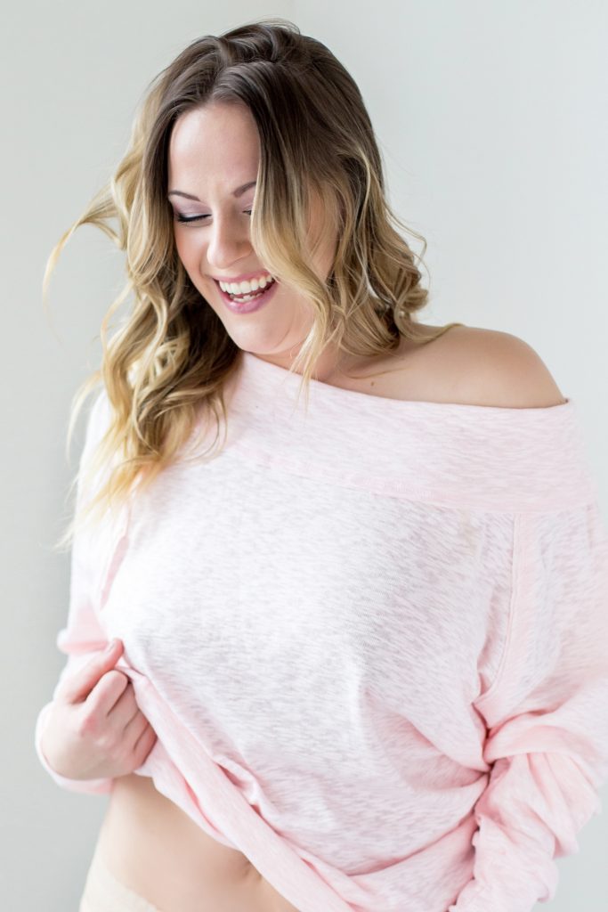 7 tips for feeling confident while being photographed. This is a modest boudoir image in a photography studio of a woman standing in front of a white background and laughing. she's wearing a pink sweater. Photography by Rayleigh. For more info, please visit byRayleigh.com
