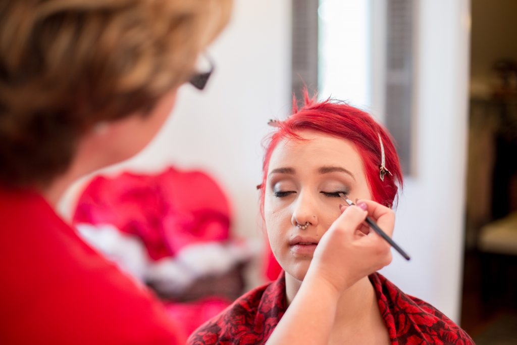 A valentine's day gift idea. Portland boudoir Photographer, Photography by Rayleigh. This woman is wearing all red and has red hair, which was a creative theme for this photo shoot. In this photo she is having her makeup done by a professional hair and makeup artist.