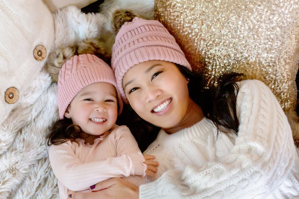 A Christmas Mother-Daughter Session - Portland Family Photographer. An Asian Mom and her young daughter lying on a bed of blankets and gold pillows, wearing pink winter hats. For more info, visit byRayleigh.com. Photography by Rayleigh