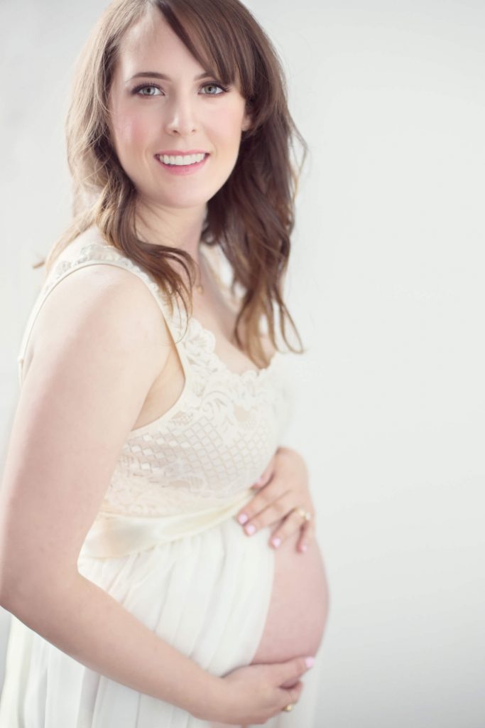 What to wear for a maternity photo shoot in Portland, OR. This is an image of a pregnant woman posing with a flowy gown in a studio with a white wall.