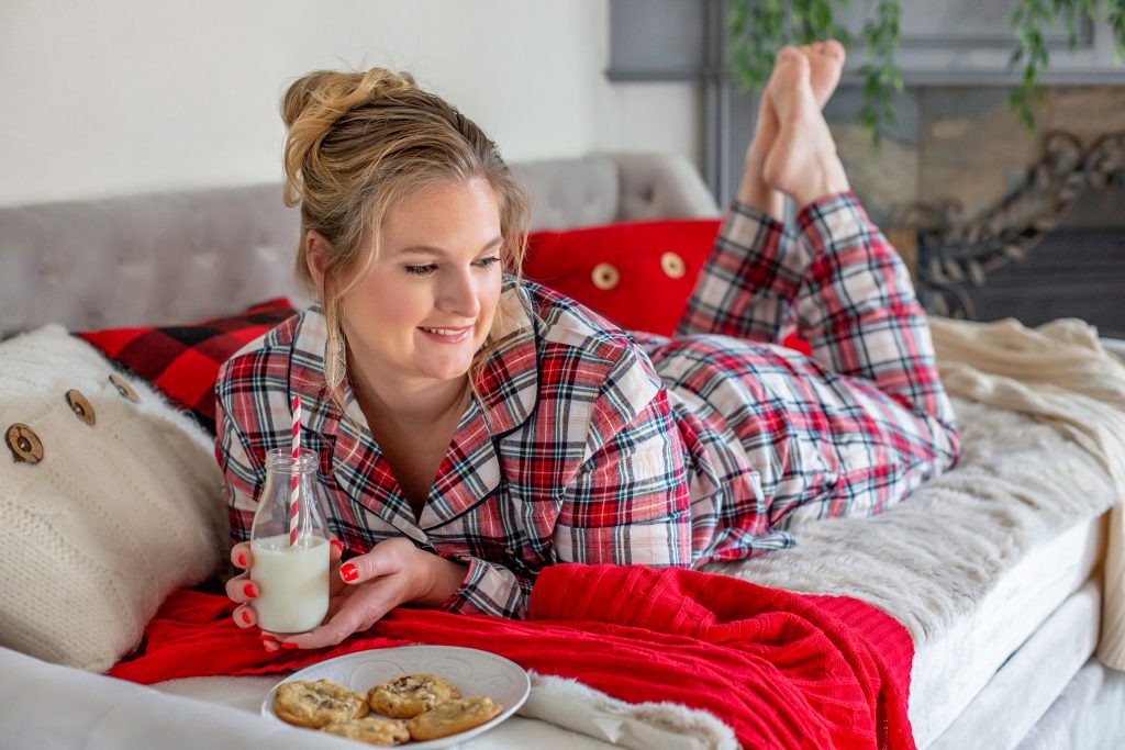 What to wear for a winter photo shoot in Portland. This is an image of a woman wearing red plaid pajamas. She's lying on her tummy on the bed with milk and cookies. Photography by Rayleigh. byRayleigh.com