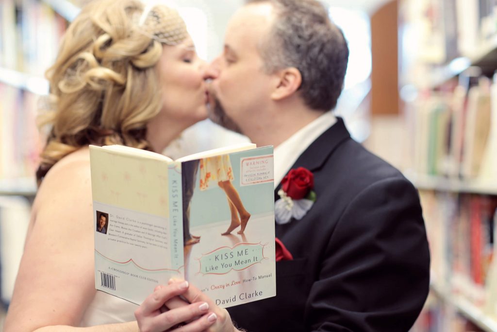 A bride and groom kissing and holding a book during their couples photo shoot at their Valentine's Day Wedding. The wedding was held at the library in Hillsboro, OR near Portland.