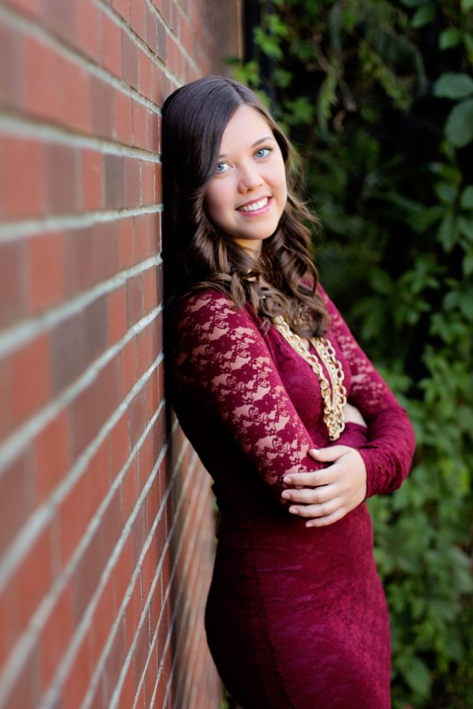 Urban senior portraits. This is an image of a high school senior girl leaning against a brick wall outdoors, wearing a red lace dress. Photography by Rayleigh. For more info, please visit byRayleigh.com