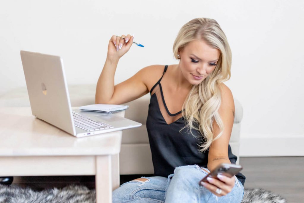 This is an image from a headshot business portrait photoshoot in Portland, OR. This branding photo shows a woman sitting on the floor and working with her laptop and cell phone while holding a pen prop.