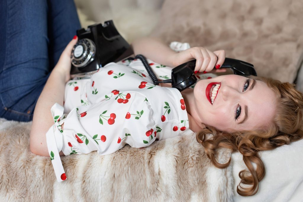What to wear for your 50's retro shoot. Portland Pin up photographer. This image is of a woman lying on her back on the bed, wearing a 50's inspired cherry button up shirt. She's holding a black rotary telephone for her prop. Photography by Rayleigh. For more info, please visit byrayleigh.com