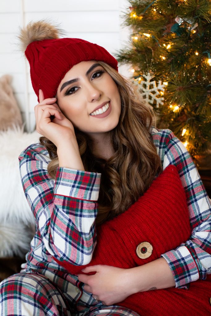 Christmas pajama sessions. This is an image of a girl wearing red plaid Christmas pajamas and a red winter hat. She's hugging a red pillow and sitting in front of a Christmas tree. Image by Photography by Rayleigh. For more info, visit byrayleigh.com