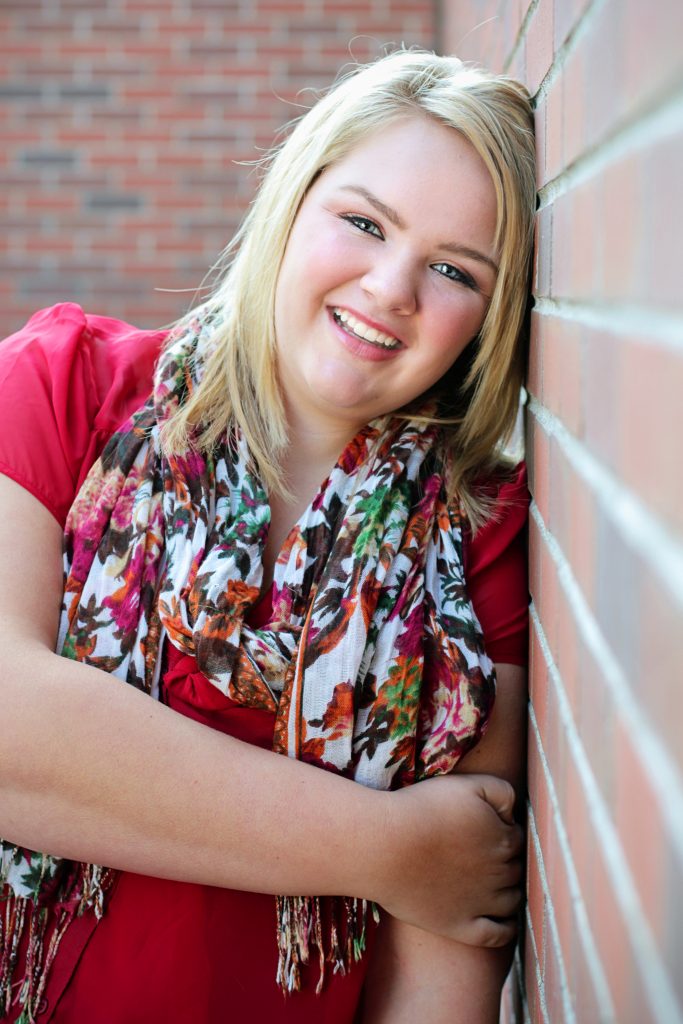 Outdoor senior pictures. This is an image of a plus size teen girl leaning against a brick wall and wearing a scarf. Photography by Rayleigh. For more info, please visit byRayleigh.com